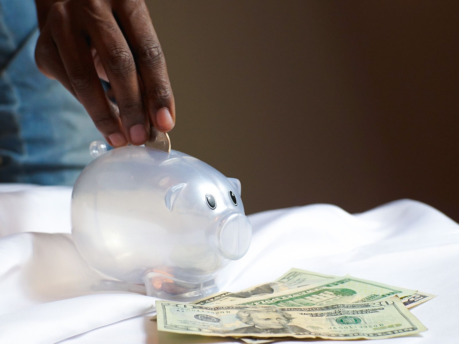 Hand placing a coin into a piggy bank beside dollar bills